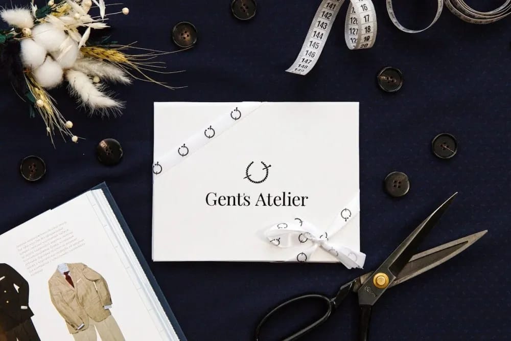 Крупный план белого конверта Gent's Atelier с фирменной лентой, ножницами и сухоцветами на синей ткани