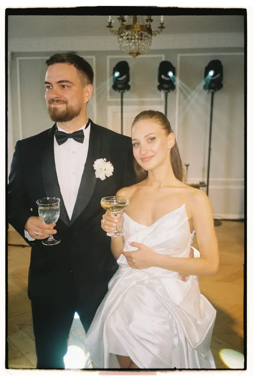Жених и невеста с бокалами шампанского на фоне зеркального зала: жених в чёрном смокинге с белой бутоньеркой, плёночный кадр.